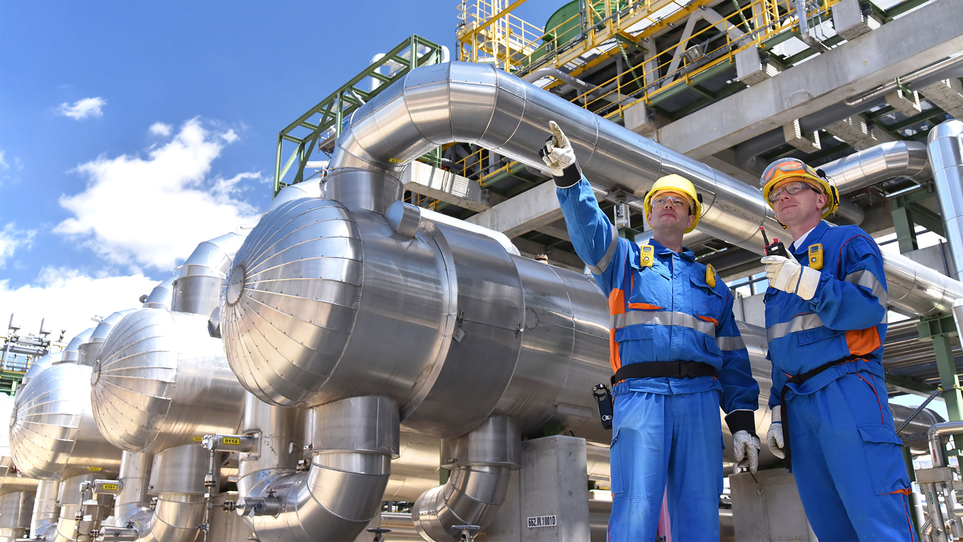 Workers in jumpsuits and protective gear standing in front of industrial chemical plant equipment.